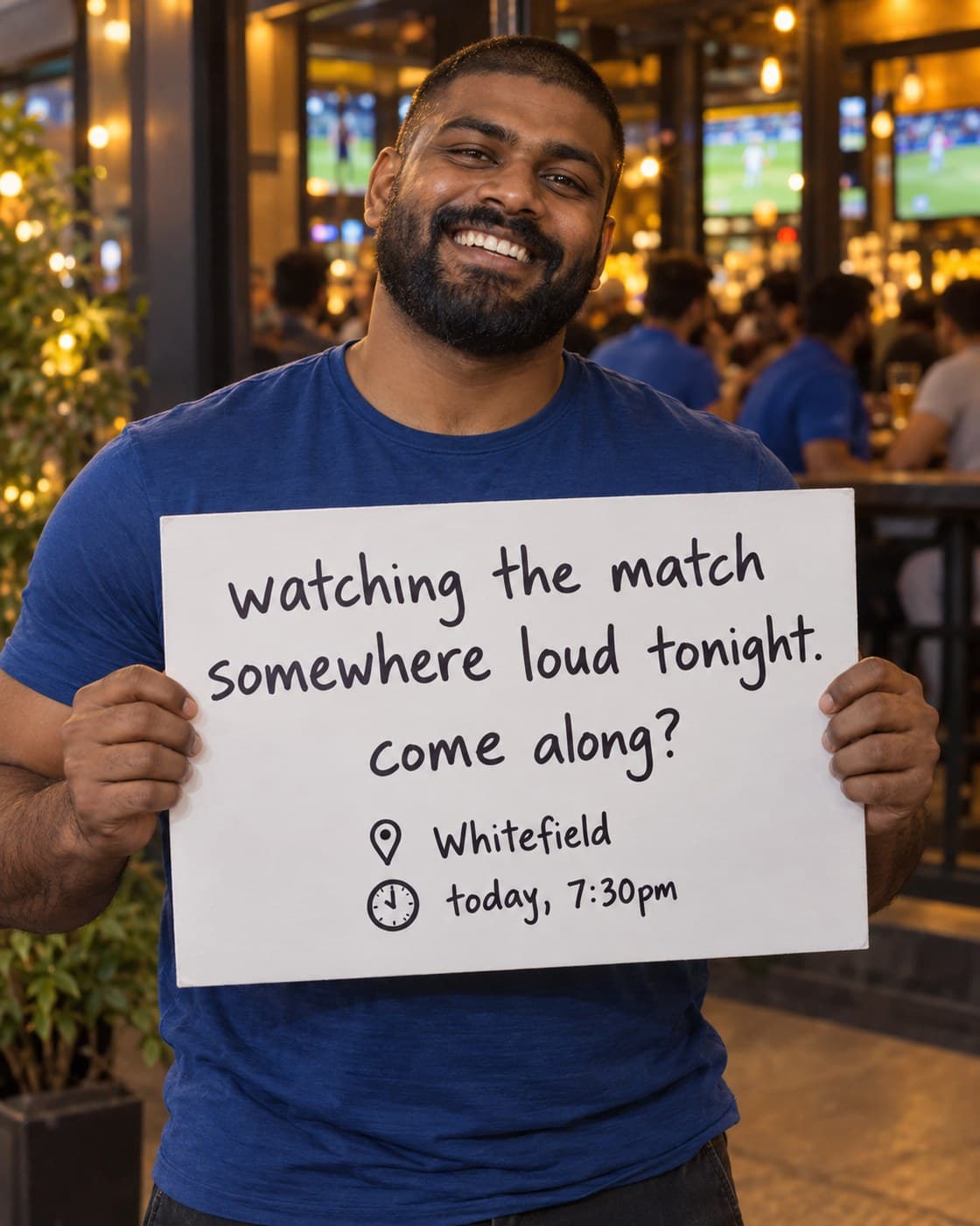 A bearded man outside a sports bar holding a sign: watching the match somewhere loud tonight. come along? Whitefield, today 7:30pm.